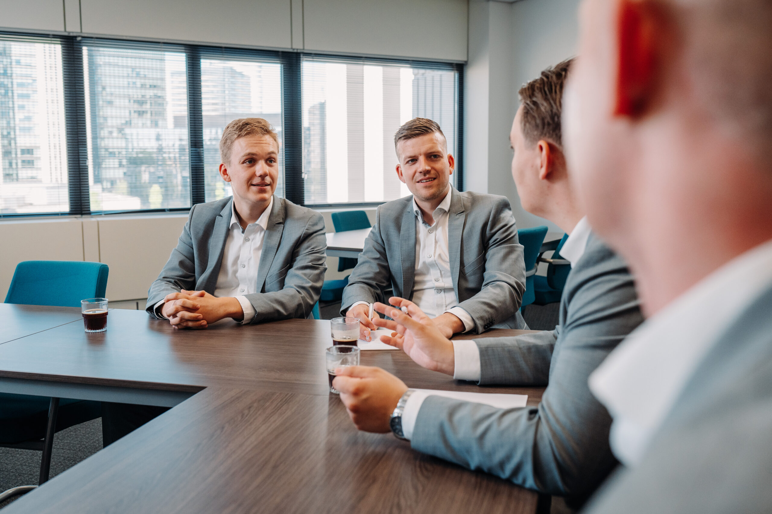 Vier mannen in pak zitten aan een vergadertafel en praten met elkaar in een moderne kantoorruimte.